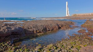 A rocky shoreline with clear water, a tall white structure, and a distant building.