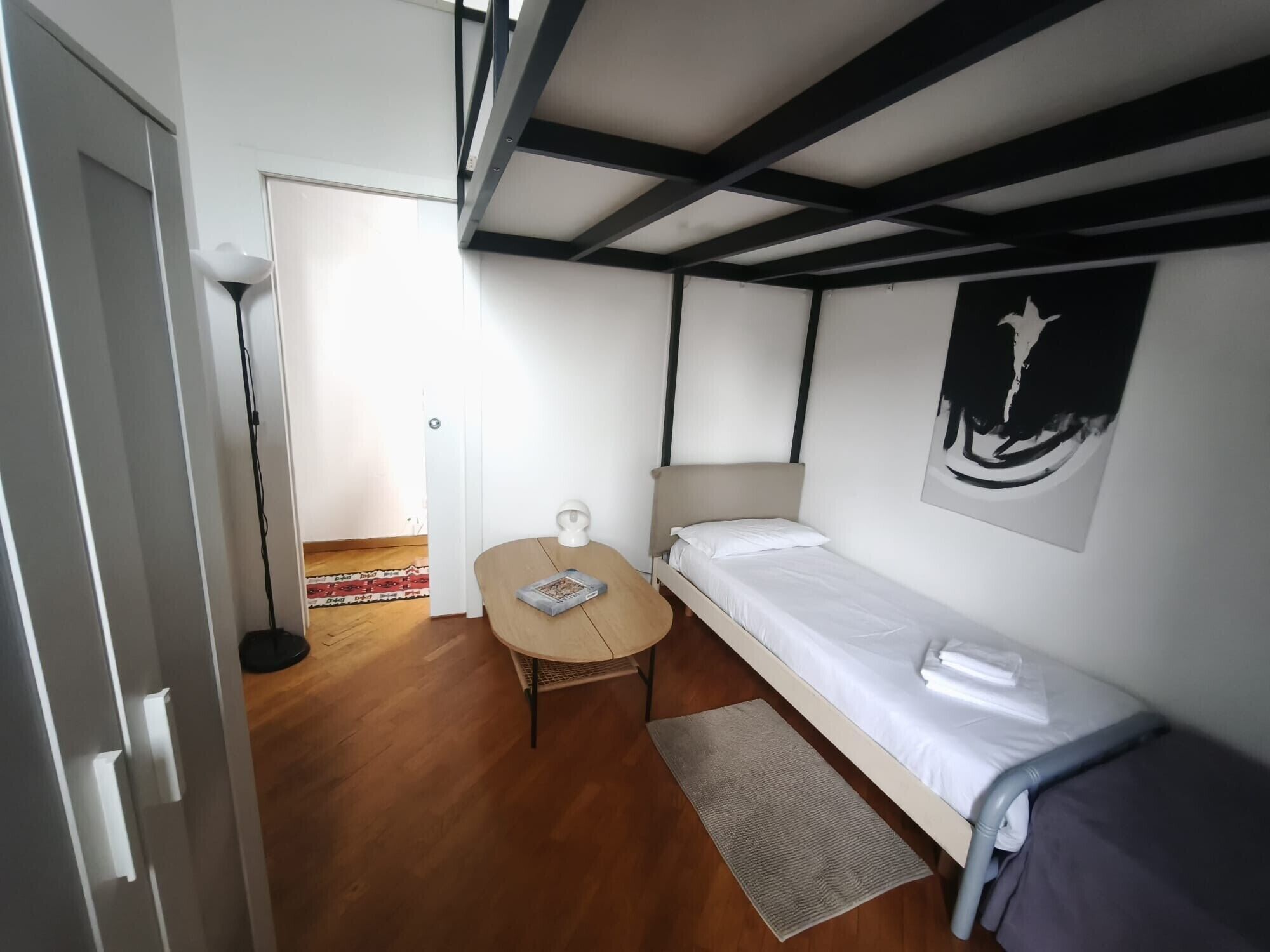 A small, single-room accommodation with a bed, a table, a chair, a lamp, and a poster on the wall.