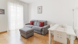 A modern living room with a grey sofa, ottoman, and white dining table with chairs.