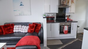 A modern living room with a black leather sofa, red throw pillows, and a white abstract painting on the wall. Adjacent is a compact kitchen with white cabinets, a red microwave, and a stainless steel oven. - I C Lundy Too (Bideford)