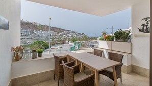 Ein Balkon mit Korbmöbeln, Blick auf eine Küstenstadt und Kübelpflanzen.