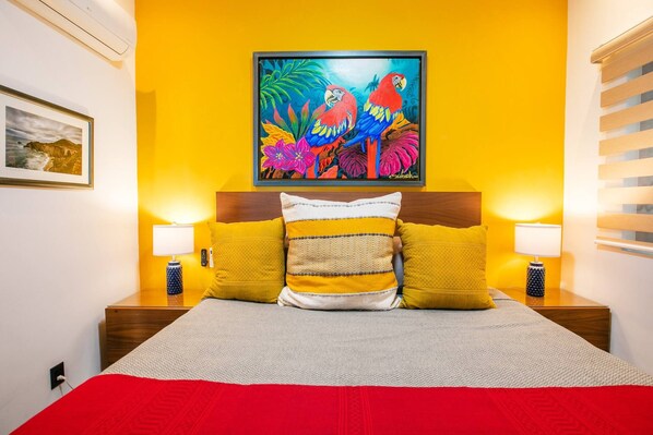 Una cama con una colcha roja, cabecera de madera y tres almohadas en tonos amarillo y blanco. Hay dos lámparas de noche de madera y un cuadro colorido de loros en la pared sobre la cama.
