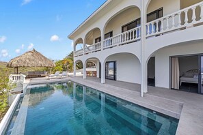Una casa grande y moderna con piscina, varios balcones y una estructura de techo de paja al fondo.