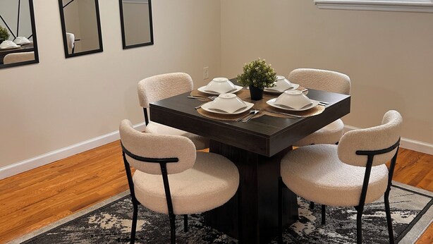 Een eetgedeelte met een donker houten tafel, witte stoelen met kussens, een zwart-wit tapijt en drie spiegels aan de muur.