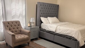 Een slaapkamer met een bed met een gestuikt hoofdeinde, een gestoffeerde fauteuil, een nachtkastje met een lamp en een grijze vloerkleed.