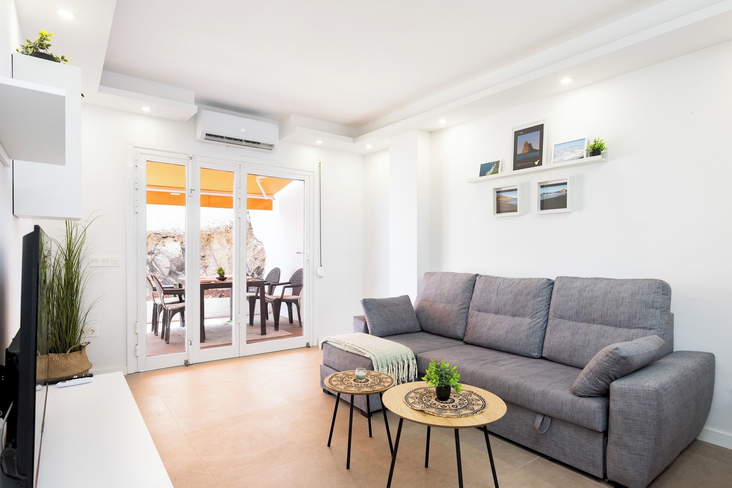 Un salon moderne avec un canapé gris, deux tables rondes ornées de plantes, un téléviseur et une porte-fenêtre coulissante donnant sur un espace extérieur.