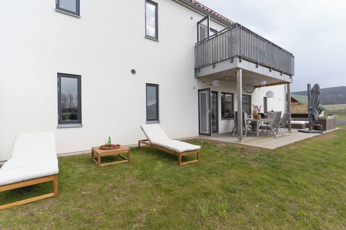 A modern house with a balcony, two lounge chairs, a table, and a dining area.
