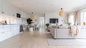 A modern kitchen with white cabinets, a dining area with a table and chairs, a sofa, and a TV mounted on the wall. - Fully Equipped Home in Bastad with Cozy Patio (Båstad)