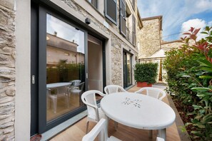 Un patio con una mesa redonda y sillas blancas, un muro de piedra y una puerta de cristal que da a un espacio interior.