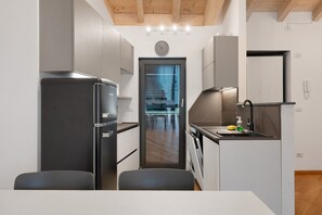 Une cuisine moderne équipée d’un réfrigérateur, d’une cuisinière et d’un évier. On y trouve une table à manger avec des chaises. Une horloge est visible au mur.