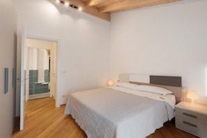 Une chambre à coucher comprenant un lit, des tables de chevet, une commode, un plafond en bois et une porte donnant sur une salle de bain.