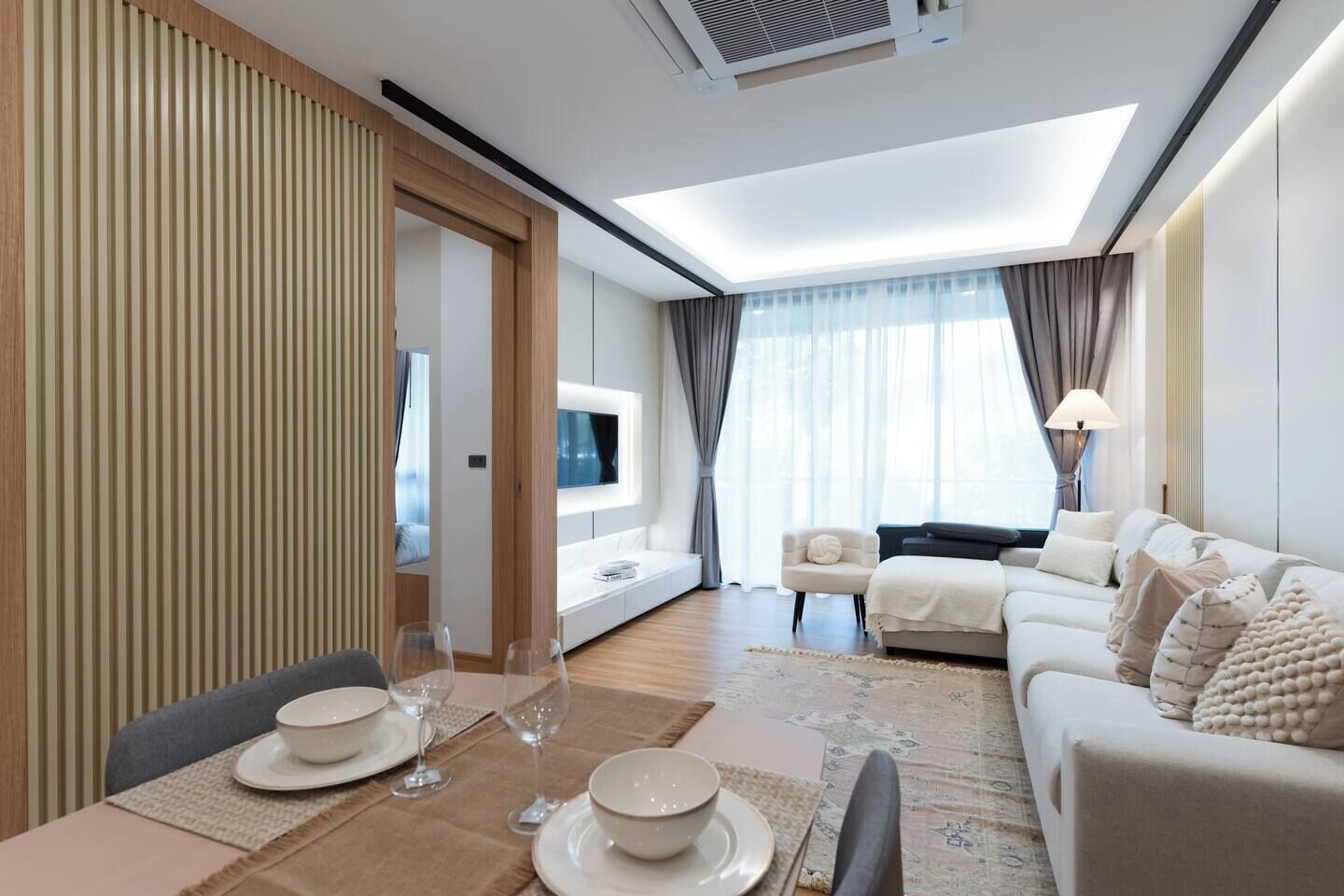 A modern hotel room with a dining area, a sofa, a TV, and a large window with curtains.