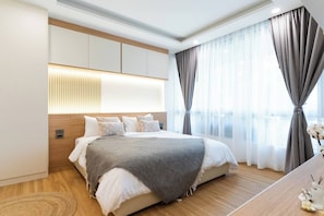 A modern bedroom with a large bed, wooden headboard, and a view of the city through sheer curtains. - Suite Coral 2br | Pool access|relife nai harn (Rawai)