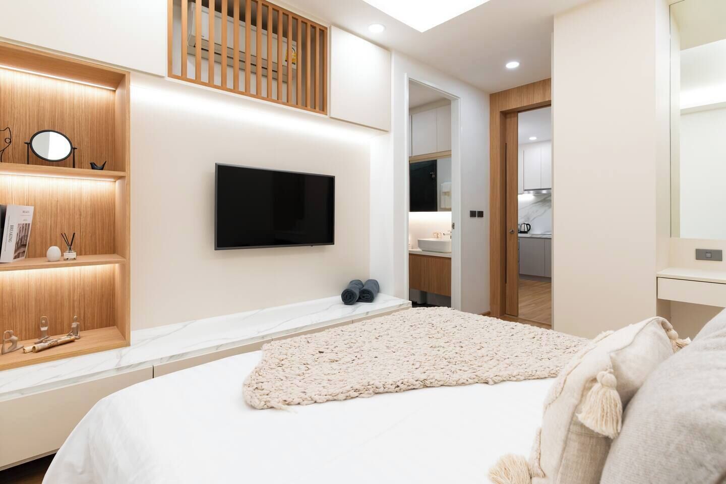 A modern bedroom with a large bed, a mounted TV, a wooden shelf with decorative items, and a view into the bathroom.