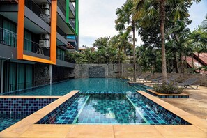 Une piscine dont les bords sont recouverts de carreaux, entourée de chaises longues et de palmiers, et située à côté d’un immeuble à plusieurs étages doté de balcons.