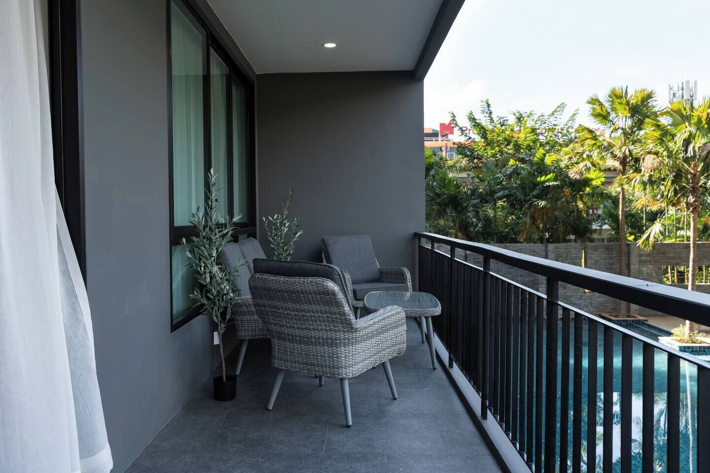 A balcony with wicker furniture, a glass table, and a view of a swimming pool and greenery.