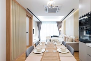 A modern dining area with a long table set for four, a sofa, and a kitchen with a built-in oven. - Suite Coral 2br | Pool access|relife nai harn (Rawai)