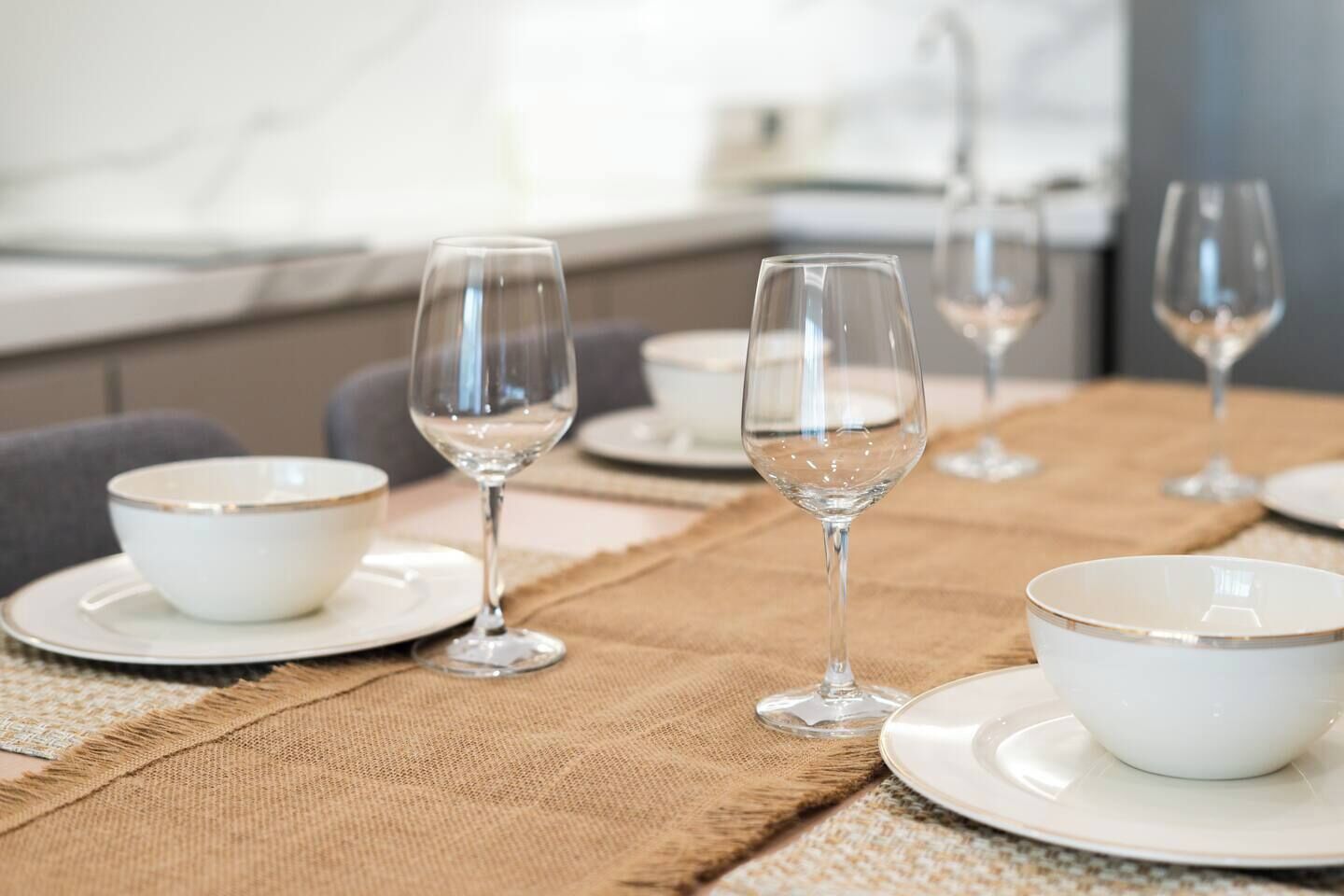 A dining table set with white plates, bowls, and clear stemmed glasses, arranged on a textured table runner.