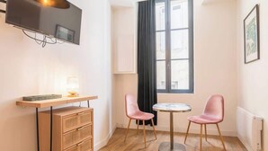 A small room with a TV mounted on the wall, a wooden cabinet, two pink chairs around a small round table, and a window with a view of buildings.