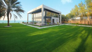 A modern building with large glass windows, a flat roof, and a well-maintained lawn with a palm tree. - Overwater Luxury Retreat - Nurai Island (Abu Dhabi)