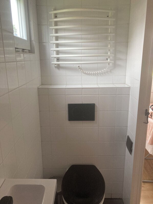 Un baño pequeño y funcional con azulejos blancos, inodoro negro, toallero y ventana.