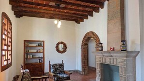 A traditional living room with exposed brick ceiling, a fireplace, a bookshelf, a floral armchair, and a patterned rug.