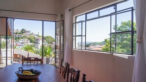 A dining area with a table, chairs, and a bowl of fruit. Large windows offer a view of buildings and greenery.