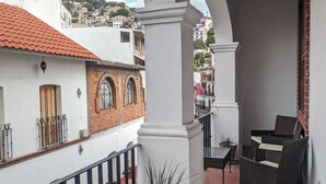 A balcony with a white railing, a potted plant, wicker furniture, and a view of a cityscape with buildings and a hill.