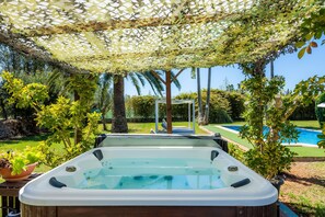 A hot tub under a pergola with a pool and greenery in the background.