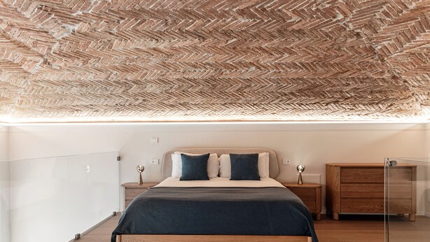 Un dormitorio moderno con techo de patrón espiga, estructura de cama de madera, mesita de noche con lámpara y cómoda de madera.