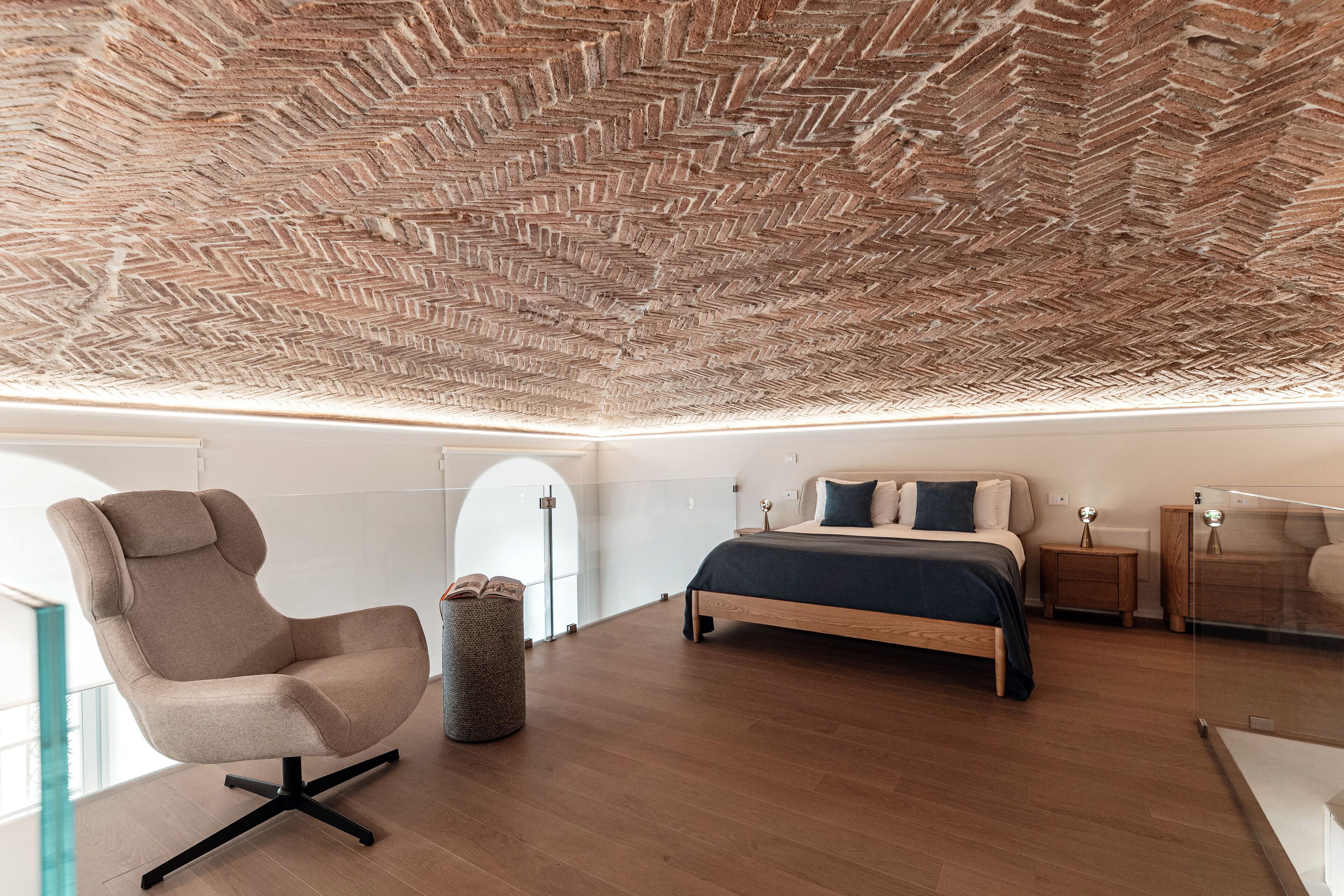 Une chambre moderne avec un plafond orné d’un motif en chevrons, un grand lit, un coin salon avec un fauteuil et une petite table de chevet.