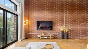 A modern living room with a brick wall, a flat-screen TV mounted above a wooden console table, and a glass coffee table. - Skyline Vibes, Walk to Stadium, Long Stay Ready (Charlotte)