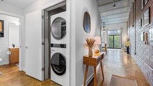 A modern laundry room with a large washing machine, a wooden table with decorative items, and a view of the outdoors through a large window. - Skyline Vibes, Walk to Stadium, Long Stay Ready (Charlotte)
