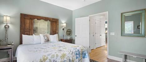 Een slaapkamer met een groot bed, een plafondventilator, een houten hoofdbord, een nachtkastje, een lamp en een deur naar een andere kamer.