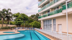 A swimming pool adjacent to a multi-story hotel with balconies, surrounded by greenery.