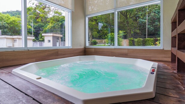 A hot tub in a room with large windows, wooden flooring, and a view of greenery outside.