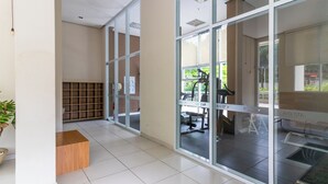 A modern hotel lobby with a tiled floor, glass doors leading to a fitness area, and a wooden bench.