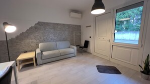 A modern living room with a grey sofa, a small table, a TV, a staircase with a grey brick wall, and a large window with a view of greenery.