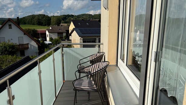 Un balcon doté d’une balustrade en verre, de deux chaises et offrant une vue sur des maisons et des collines.