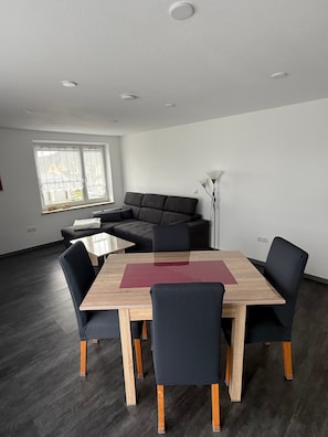 Ein modernes Wohnzimmer mit einem dunkelgrauen Sofa, einem Holzesstisch mit vier Stühlen und einer Stehlampe.