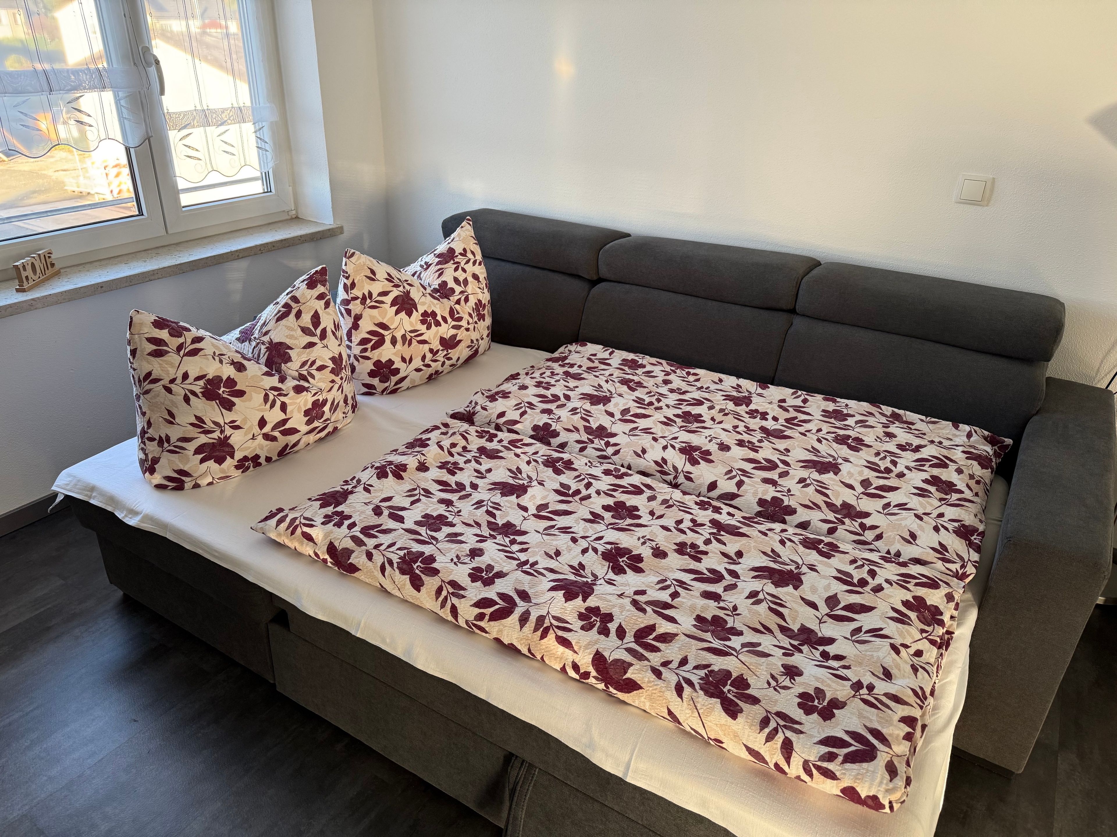 Una cama con colcha estampada, dos almohadas y una mesita de noche en una habitación con ventana.