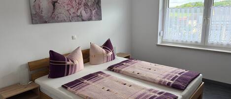A bedroom with a bed, two pillows, a nightstand, a drawer, a window with blinds, and a floral painting on the wall.