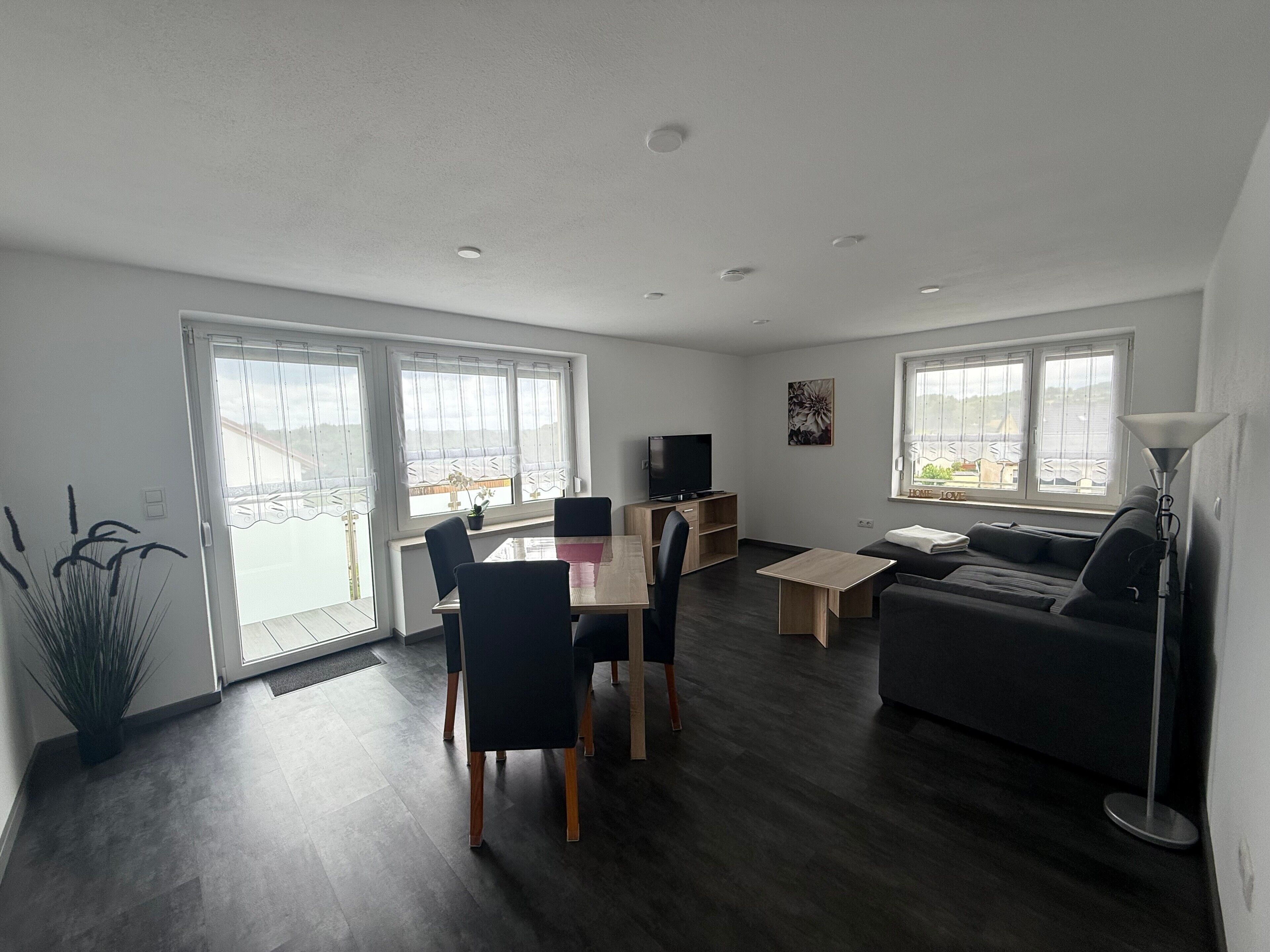 A modern living room with a dining area, a sofa, a TV, and large windows.