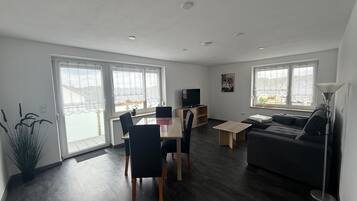 A modern living room with a dining area, a sofa, a TV, and large windows.