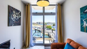 A living room with a sofa, a painting, and a view of a pool and cityscape through a large window. - Cosy Apartment with Valletta and Harbour Views (Sliema)