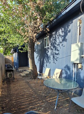 Ein Garten mit Baum, Tisch und Stühlen. Im Hintergrund ist ein blaues Gebäude zu sehen.
