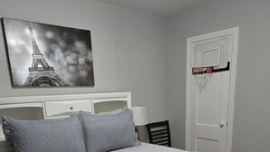 Une chambre avec un lit, un ventilateur de plafond, une porte sur laquelle est fixé un panier de basket et un tableau représentant la Tour Eiffel, accroché au mur.