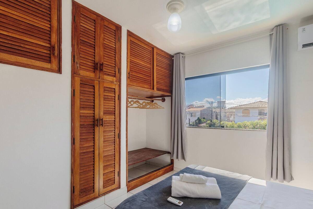 Une pièce avec des volets en bois, un lit avec un matelas bleu, une fenêtre donnant sur des immeubles et un luminaire au plafond.
