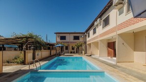 A swimming pool with a pergola and palm trees, a building with a tiled roof, and a clear blue sky. - Cond. c\ Piscina e Churrasqueira a 650 m Praia de Taperapuan \ PFNs (Porto Seguro)