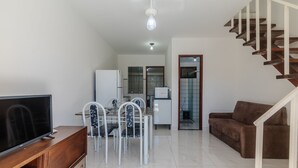A living room with a TV, dining table, chairs, a sofa, and a staircase. - Cond. c\ Piscina e Churrasqueira a 650 m Praia de Taperapuan \ PFNs (Porto Seguro)